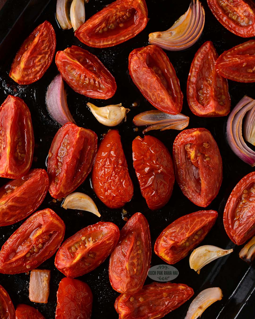 Oven roasted tomatoes garlic cloves onion on a black baking tray.